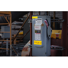A piece of electrical equipment is attached to a beam indoors. It is marked with a number of labels. 