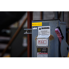 A piece of electrical equipment is attached to a beam indoors. It is marked with a number of labels. 
