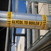 A pipe in an outdoor setting with a yellow pipe marker that reads “Glycol to Boiler 2" and a set of wraparound arrows on each end.