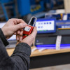 A close-up of a person's hands applying a label to a red safety padlock. The background is blurred, showing a label printer and other industrial equipment.