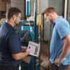 Two people are standing in front of industrial machinery and piping. The person on the left is holding a lockout procedure and pointing to it to instruct the person on the right.