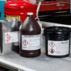 Three containers on a table in an industrial facility, each with a GHS label applied.
