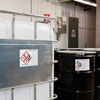 A large white container encase by metal bars and a large black drum in an industrial facility, each with a GHS label applied.